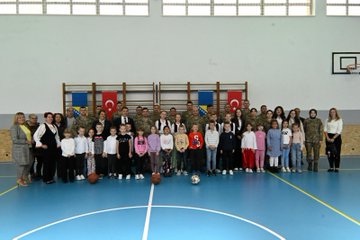 First image shows a group of children in black tops and colorful skirts including red purple and blue holding hands in a circle on a gymnasium floor with Turkish flags and a banner reading BOSNA HERSEK TÜRK TEMSİL HEYET BAŞKANLIĞI behind them. Second image displays boys wearing red fezzes and white traditional outfits with red sashes kneeling and performing on a stage flanked by banners for BOSNA HERSEK TÜRK TEMSİL HEYET BAŞKANLIĞI and VRSKA MUJA and girls in headscarves and white dresses beside them. Third image captures a large group of children in casual clothes and adults including women in professional attire and men in military uniforms posing together on a blue basketball court with Turkish and Bosnian flags on the walls and a basketball on the floor. Fourth image features another group photo of children holding small Turkish flags some in pink outfits and adults in military camouflage and civilian clothes standing in a hallway with Bosnian flags and educational posters visible.