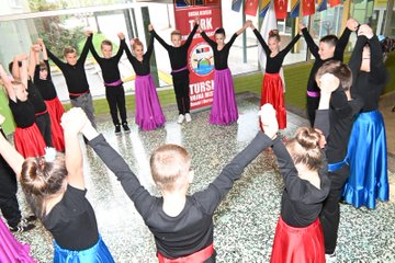 First image shows a group of children in black tops and colorful skirts including red purple and blue holding hands in a circle on a gymnasium floor with Turkish flags and a banner reading BOSNA HERSEK TÜRK TEMSİL HEYET BAŞKANLIĞI behind them. Second image displays boys wearing red fezzes and white traditional outfits with red sashes kneeling and performing on a stage flanked by banners for BOSNA HERSEK TÜRK TEMSİL HEYET BAŞKANLIĞI and VRSKA MUJA and girls in headscarves and white dresses beside them. Third image captures a large group of children in casual clothes and adults including women in professional attire and men in military uniforms posing together on a blue basketball court with Turkish and Bosnian flags on the walls and a basketball on the floor. Fourth image features another group photo of children holding small Turkish flags some in pink outfits and adults in military camouflage and civilian clothes standing in a hallway with Bosnian flags and educational posters visible.