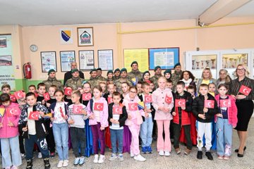 First image shows a group of children in black tops and colorful skirts including red purple and blue holding hands in a circle on a gymnasium floor with Turkish flags and a banner reading BOSNA HERSEK TÜRK TEMSİL HEYET BAŞKANLIĞI behind them. Second image displays boys wearing red fezzes and white traditional outfits with red sashes kneeling and performing on a stage flanked by banners for BOSNA HERSEK TÜRK TEMSİL HEYET BAŞKANLIĞI and VRSKA MUJA and girls in headscarves and white dresses beside them. Third image captures a large group of children in casual clothes and adults including women in professional attire and men in military uniforms posing together on a blue basketball court with Turkish and Bosnian flags on the walls and a basketball on the floor. Fourth image features another group photo of children holding small Turkish flags some in pink outfits and adults in military camouflage and civilian clothes standing in a hallway with Bosnian flags and educational posters visible.