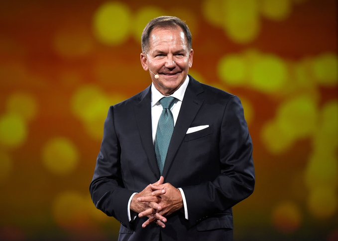 A man in a dark suit with green tie stands on stage with hands clasped, smiling, against a blurred orange and yellow background with spotlight effects. A white Bitcoin symbol appears on an orange background.