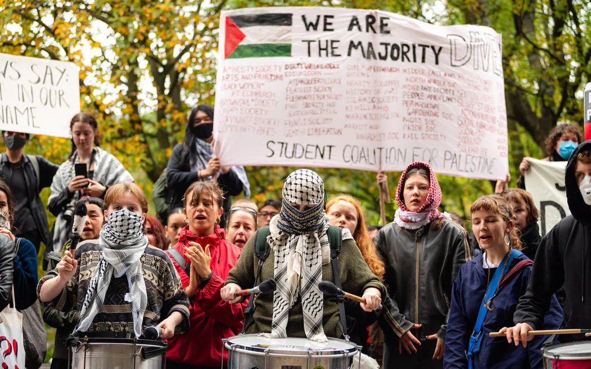 Group of young people gathered outdoors in autumn foliage, holding large white banner with black text We are the Majority and Palestinian flag emblem, another banner reading Student Coalition for Palestine with additional text, several participants wearing keffiyeh scarves and masks, some playing drums including a central figure in black keffiyeh and jacket striking a drum, others clapping and gesturing excitedly, diverse clothing like hoodies and coats, Palestinian flags visible.