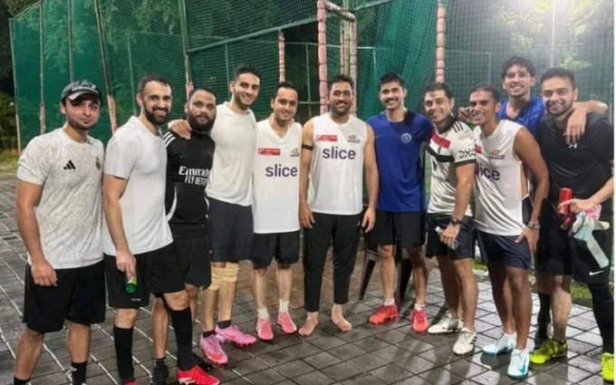 Group of men standing together on a wet outdoor surface near green netting, wearing athletic clothing including tank tops and shorts from brands like Emirates, Slice, and Puma. MS Dhoni is in the center wearing a white Mumbai Indians tank top, surrounded by players in various team jerseys and casual sportswear. Some hold water bottles, and they pose with arms around each other in a casual post-game setting.