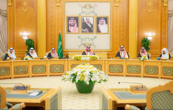 First image shows a man in traditional Saudi attire including red and white checkered headscarf and black agal, white thobe with gold trim, seated at a wooden desk with papers and green boxes, Saudi flag in background, yellow and white flowers on desk. Second image depicts the same man in similar attire, focused closely while looking down at documents on a table with flowers. Third image displays multiple men in traditional white thobes and headscarves seated around a large oval wooden table in an ornate room with gold decorations, Saudi flags, portraits on walls, central green vase with white and yellow flowers. Fourth image shows a similar group of men in formal attire around a round wooden table in a grand hall with chandeliers, draped walls, portraits, Saudi flags, and a central vase of flowers.