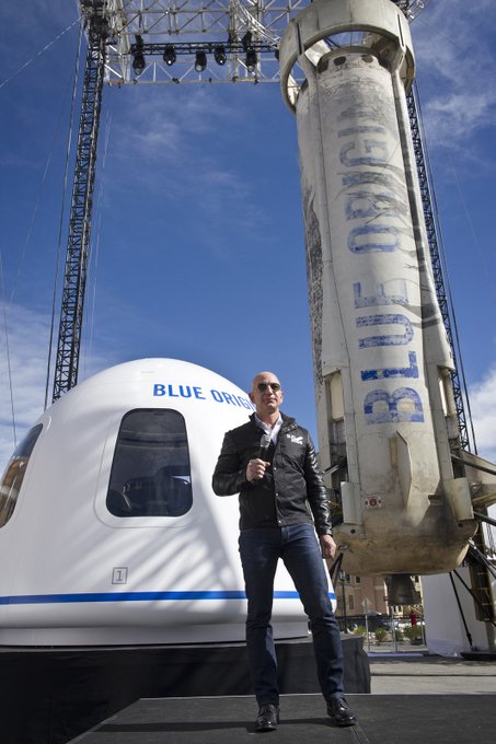 Man in leather jacket and sunglasses stands on stage holding microphone next to white Blue Origin spacecraft module with Blue Origin text on side large white Blue Origin rocket with Blue Origin lettering stands behind under blue sky with stage structure and lights visible