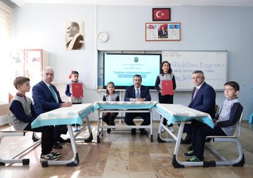 First image shows a group of schoolchildren and adults seated around two blue-covered tables with corn on them in a classroom with Turkish flags, a clock, and a smartboard displaying a ministry logo, students holding documents and smiling. Second image depicts similar group with men in suits and children holding red folders, tables covered in blue cloth, classroom setting with flags and educational posters. Third image features a man in a suit signing a document at a desk with two children standing nearby, whiteboard with Turkish text about school life and Erzurum, date 10/12/05 visible. Fourth image captures a man in a suit hugging a child while surrounded by a group of smiling schoolchildren in uniforms, table with a green bag and documents, smartboard showing Saglikla Gulums text.