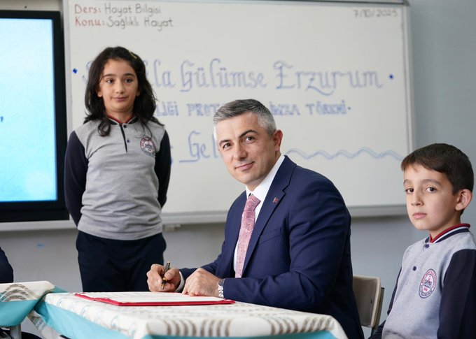 First image shows a man in a blue suit and red tie sitting at a desk in a classroom signing a document with a pen while two young children in school uniforms stand nearby one girl with long hair in a gray shirt and black pants and one boy in a gray shirt and blue pants. A whiteboard behind displays Turkish text about the school year starting in Erzurum and a screen shows presentation slides. The setting includes desks chairs and educational materials. Second image depicts a group of schoolchildren in uniforms and two adult men one in a suit holding documents with corn cobs on blue tables in a classroom with Turkish flags a clock and a portrait on the wall behind them. The children are smiling and gathered around the tables in what appears to be a celebratory educational event.