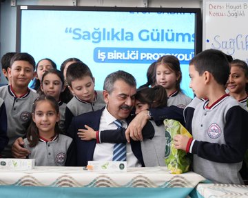 First image shows a group of schoolchildren and adults seated around two blue-covered tables with corn on them in a classroom with Turkish flags, a clock, and a smartboard displaying a ministry logo, students holding documents and smiling. Second image depicts similar group with men in suits and children holding red folders, tables covered in blue cloth, classroom setting with flags and educational posters. Third image features a man in a suit signing a document at a desk with two children standing nearby, whiteboard with Turkish text about school life and Erzurum, date 10/12/05 visible. Fourth image captures a man in a suit hugging a child while surrounded by a group of smiling schoolchildren in uniforms, table with a green bag and documents, smartboard showing Saglikla Gulums text.