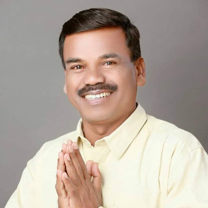A man with short dark hair and a mustache smiles while pressing his palms together in a greeting gesture, wearing a light yellow collared shirt against a plain gray background.