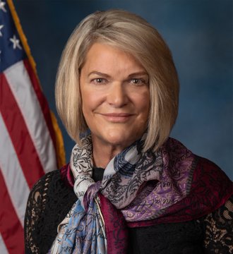 Professional headshot of Senator Cynthia Lummis, a woman with blonde bob haircut, smiling, wearing patterned scarf over black top, standing before blue background with US flag on right side. Second image displays white Bitcoin symbol on solid orange background.