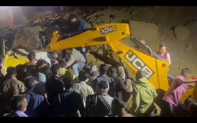 Yellow JCB excavator with black JCB logo digs through large pile of brown soil and rocks at night under orange lighting with several people in casual clothes including blue shirts jeans and hoodies gathered around observing the operation some standing on the debris.