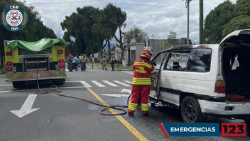 First image shows a firefighter in red protective gear and helmet spraying water from a high-pressure hose at a white van with severe fire damage on its side and roof inside a cemetery surrounded by tombstones and a low wall with the Bomberos Municipales emblem visible. Second image depicts another firefighter in similar gear examining the open rear of the same white van on a street with trees and people in the background while a green fire truck is connected to a hose nearby. Third image features a firefighter in red gear with helmet directing a hose into the open side door of the burned white van on a road with a green fire truck and traffic signs in the view.