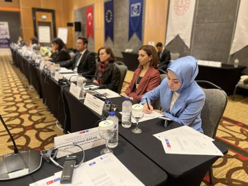 First image shows a wooden podium with a laptop displaying a presentation slide titled in Turkish about foreign language education project, microphone, water bottle, and pen on a carpeted stage in a conference room with wooden walls, EU and Turkish flags, and banners. Second image depicts a large oval conference table with multiple participants in business attire seated around it, a large screen projecting a slide, wooden paneled walls, chandeliers, and flags including Turkish and EU banners in a grand meeting hall. Third image captures a close-up of six professionals including men in suits and a woman in a blouse seated at a black conference table with name cards, water bottles, documents, and microphones, focused on discussion. Fourth image features a row of diverse participants including women in headscarves and men in suits at a long black table with papers, glasses of water, microphones, and name tags like Mahmut Ozdil, against a backdrop of Turkish and EU flags in a formal setting.