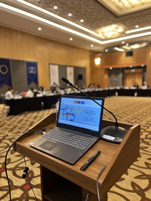 First image shows a wooden podium with a laptop displaying a presentation slide titled in Turkish about foreign language education project, microphone, water bottle, and pen on a carpeted stage in a conference room with wooden walls, EU and Turkish flags, and banners. Second image depicts a large oval conference table with multiple participants in business attire seated around it, a large screen projecting a slide, wooden paneled walls, chandeliers, and flags including Turkish and EU banners in a grand meeting hall. Third image captures a close-up of six professionals including men in suits and a woman in a blouse seated at a black conference table with name cards, water bottles, documents, and microphones, focused on discussion. Fourth image features a row of diverse participants including women in headscarves and men in suits at a long black table with papers, glasses of water, microphones, and name tags like Mahmut Ozdil, against a backdrop of Turkish and EU flags in a formal setting.