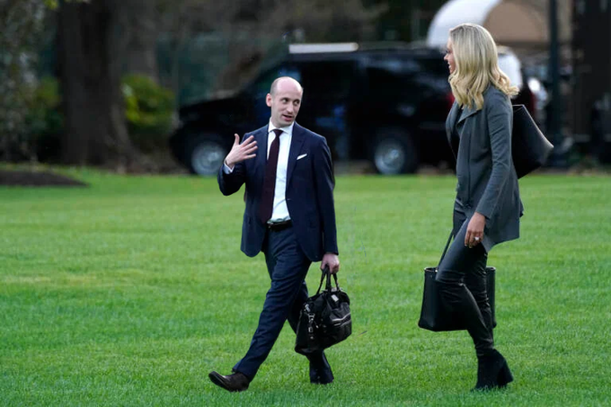 A bald man in a dark suit with a red tie, carrying a black handbag, walks on green grass while gesturing with his right hand raised. Beside him is a blonde woman in a gray coat, black pants, and knee-high black boots, holding a black handbag. A black SUV is parked nearby with an open door, trees and another vehicle in the background.