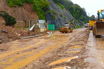 Aerial view of a highway section with a large landslide covering the road, surrounded by green vegetation and a body of water nearby, showing excavators and dump trucks positioned for cleanup. Ground-level shot of yellow excavators and bulldozers working on muddy terrain beside a sloped embankment with debris, workers in safety gear operating machinery, and barriers in place. Side view along a wet road with multiple yellow heavy machines like excavators clearing earth and rocks from the roadside, people walking nearby, and a billboard visible in the background. Close-up of a blue and yellow John Deere backhoe loader with visible branding, digging into muddy soil near another excavator, all on a debris-strewn path with overhanging trees.