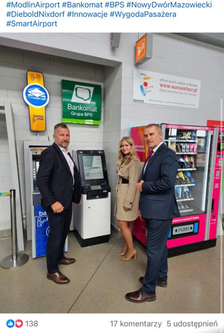 Three individuals stand smiling in a modern airport terminal: a man in a dark suit and brown shoes on the left, a woman in a beige trench coat and heels in the center, and another man in a gray suit and brown shoes on the right. They pose in front of a white Diebold Nixdorf ATM machine displaying Bank Pekao branding. To the right is a pink vending machine stocked with snacks. Green and yellow signs for BPS Group and Nowy Dwór Mazowiecki are mounted on walls, along with directional and safety signs. The floor is tiled, and overhead lighting illuminates the area.