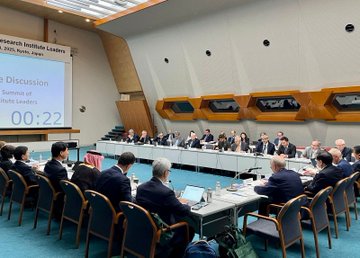 First image shows a large conference room with a long rectangular table surrounded by chairs occupied by numerous professionals in business attire engaged in a meeting with laptops documents and a projector stand in the center on a blue carpeted floor with modern architectural elements like wooden panels and large windows. Second image depicts Prof. Dr. Orhan Aydın a middle-aged man with gray hair wearing glasses a dark suit a green tie and a name tag labeled Orhan Aydın TÜBİTAK Türkiye seated at a table with a microphone water bottle papers and a red folder in front of him in a meeting setting. Third image displays a conference room with a screen showing Institute Leaders Summit Discussion in English and Japanese with a timer at 00:22 and a group of international professionals in suits seated around a U-shaped table with laptops documents and water bottles on a blue carpeted floor under modern architecture. Fourth image features a large group photo of diverse international leaders including men and women in formal suits some with traditional attire like a thobe standing and sitting in rows against a plain backdrop in a hall with blue flooring.