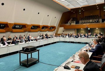 First image shows a large conference room with a long rectangular table surrounded by chairs occupied by numerous professionals in business attire engaged in a meeting with laptops documents and a projector stand in the center on a blue carpeted floor with modern architectural elements like wooden panels and large windows. Second image depicts Prof. Dr. Orhan Aydın a middle-aged man with gray hair wearing glasses a dark suit a green tie and a name tag labeled Orhan Aydın TÜBİTAK Türkiye seated at a table with a microphone water bottle papers and a red folder in front of him in a meeting setting. Third image displays a conference room with a screen showing Institute Leaders Summit Discussion in English and Japanese with a timer at 00:22 and a group of international professionals in suits seated around a U-shaped table with laptops documents and water bottles on a blue carpeted floor under modern architecture. Fourth image features a large group photo of diverse international leaders including men and women in formal suits some with traditional attire like a thobe standing and sitting in rows against a plain backdrop in a hall with blue flooring.