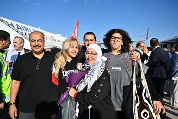 First image shows a bearded man with long hair smiling while embracing two young boys, one holding a bouquet of red roses, surrounded by a crowd of people in traditional attire at an indoor event with bright lights. Second image depicts a woman in a pink top and headscarf smiling while hugging a man in black, holding a phone taking a selfie, with other women in hijabs and a diverse group in the background under event lighting. Third image features a group of five people posing in front of a Turkish Airlines airplane on a tarmac under blue sky, including a blonde woman in black, a woman in hijab holding purple flowers, a young man with glasses, and two men in dark shirts, with Turkish and Palestinian flags visible. Fourth image captures a large group of men and women, some in Palestinian keffiyehs, standing around a podium with microphones during a formal indoor event, Turkish flags and portraits on walls, including children and media equipment.