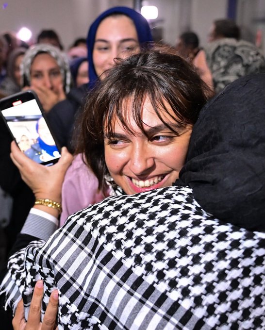 First image shows a bearded man with long hair smiling while embracing two young boys, one holding a bouquet of red roses, surrounded by a crowd of people in traditional attire at an indoor event with bright lights. Second image depicts a woman in a pink top and headscarf smiling while hugging a man in black, holding a phone taking a selfie, with other women in hijabs and a diverse group in the background under event lighting. Third image features a group of five people posing in front of a Turkish Airlines airplane on a tarmac under blue sky, including a blonde woman in black, a woman in hijab holding purple flowers, a young man with glasses, and two men in dark shirts, with Turkish and Palestinian flags visible. Fourth image captures a large group of men and women, some in Palestinian keffiyehs, standing around a podium with microphones during a formal indoor event, Turkish flags and portraits on walls, including children and media equipment.