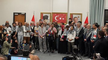 First image shows a bearded man with long hair smiling while embracing two young boys, one holding a bouquet of red roses, surrounded by a crowd of people in traditional attire at an indoor event with bright lights. Second image depicts a woman in a pink top and headscarf smiling while hugging a man in black, holding a phone taking a selfie, with other women in hijabs and a diverse group in the background under event lighting. Third image features a group of five people posing in front of a Turkish Airlines airplane on a tarmac under blue sky, including a blonde woman in black, a woman in hijab holding purple flowers, a young man with glasses, and two men in dark shirts, with Turkish and Palestinian flags visible. Fourth image captures a large group of men and women, some in Palestinian keffiyehs, standing around a podium with microphones during a formal indoor event, Turkish flags and portraits on walls, including children and media equipment.