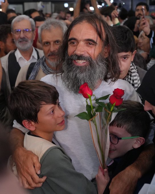 First image shows a bearded man with long hair smiling while embracing two young boys, one holding a bouquet of red roses, surrounded by a crowd of people in traditional attire at an indoor event with bright lights. Second image depicts a woman in a pink top and headscarf smiling while hugging a man in black, holding a phone taking a selfie, with other women in hijabs and a diverse group in the background under event lighting. Third image features a group of five people posing in front of a Turkish Airlines airplane on a tarmac under blue sky, including a blonde woman in black, a woman in hijab holding purple flowers, a young man with glasses, and two men in dark shirts, with Turkish and Palestinian flags visible. Fourth image captures a large group of men and women, some in Palestinian keffiyehs, standing around a podium with microphones during a formal indoor event, Turkish flags and portraits on walls, including children and media equipment.