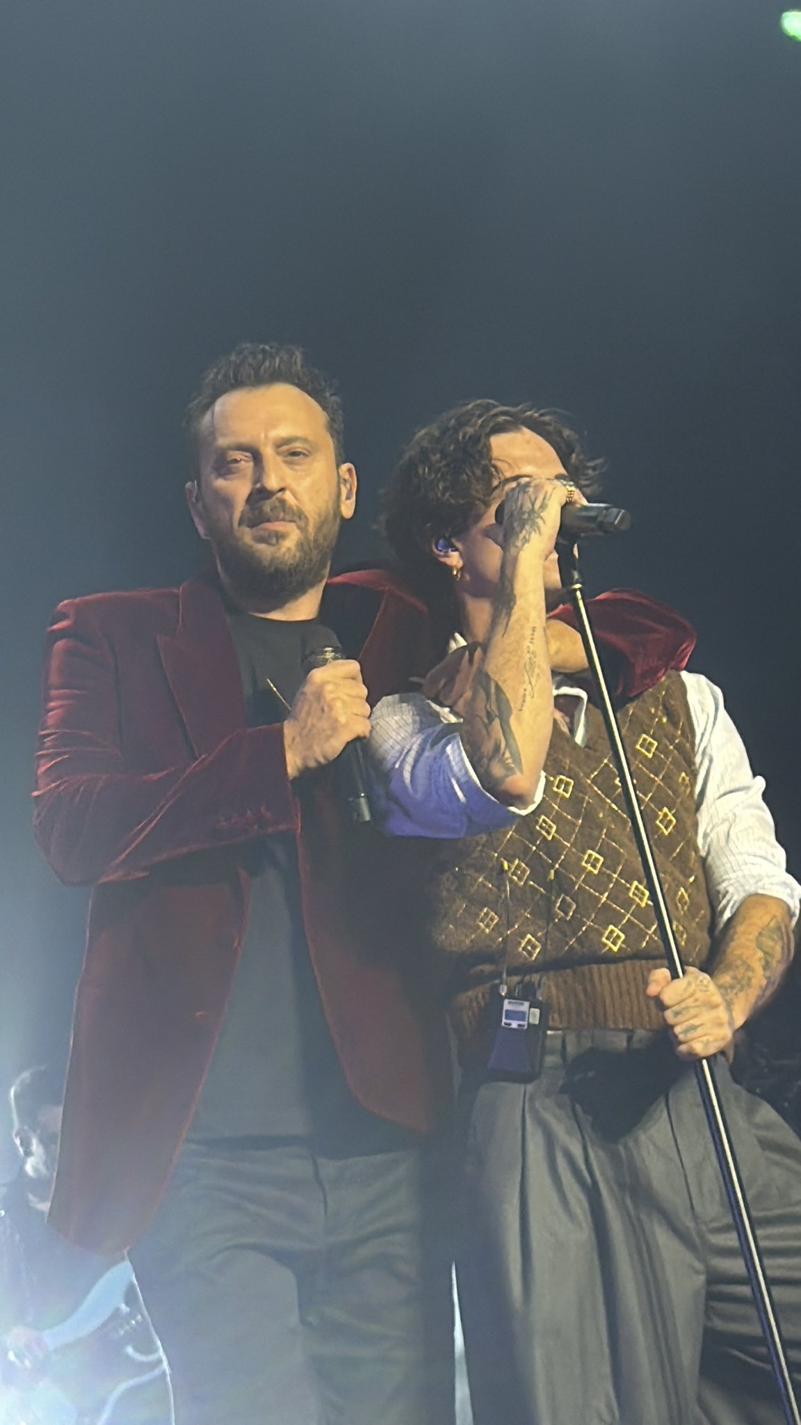 Damiano David with curly dark hair wears a red velvet blazer over a black shirt and holds a microphone near his mouth while gesturing with his left hand. Cesare Cremonini with wavy dark hair stands next to him wearing a brown Louis Vuitton monogram vest over a white shirt and gray pants, holding a microphone in his right hand and gesturing with his left. Both are on a dark stage illuminated by green and blue spotlights from above.