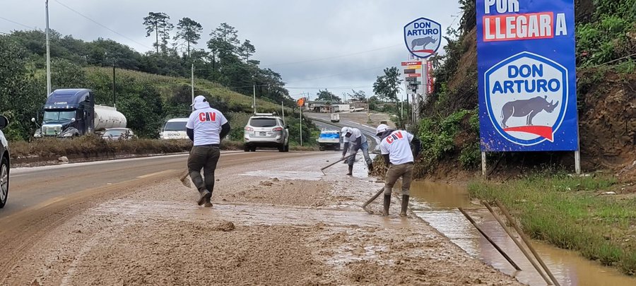 First image displays several workers in yellow vests and red shirts using shovels to clear mud and debris from a roadside area affected by a landslide, with orange traffic cones, a parked SUV, and a dirt embankment in the background under a partly cloudy sky. Second image shows workers in white shirts with red lettering and blue pants clearing wet mud from a highway shoulder near a sign reading PODER PARA LEGAR A and featuring a blue emblem, with trucks, a white vehicle, and green hills visible along the road.
