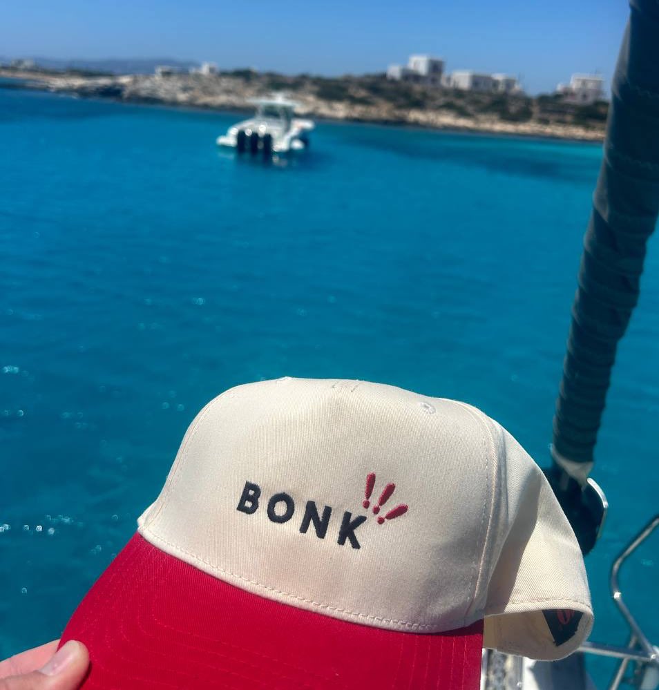 Bonk Inu hat with 'BONK!!' embroidery held up against a turquoise sea and boat