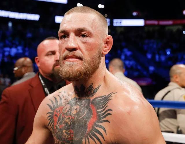 Conor McGregor está de pie sin camisa en un escenario de arena con poca luz, mostrando intrincados tatuajes en el pecho y los brazos, incluido un gran diseño central con alas y un motivo de calavera. Tiene la cabeza rapada, barba y expresión concentrada mientras usa pantalones cortos de lucha. Las personas que rodean con trajes y atuendos de seguridad son visibles en el fondo borroso cerca de un área de ring.