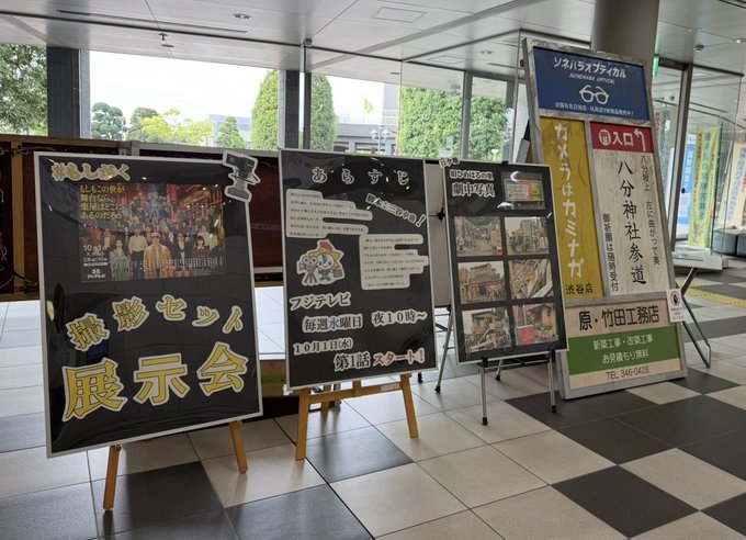 Several standing display boards and posters set up in a spacious indoor lobby with large windows showing greenery outside and tiled floors. One board features a black background with multiple people in formal attire gathered around a central figure. Another shows a black and white poster with Japanese text and images of scenes. A yellow board displays large text announcing an event with numbers like 100th anniversary. Additional panels include photo collages of urban and indoor settings with overlaid text. Easels and stands support the exhibits near walls.