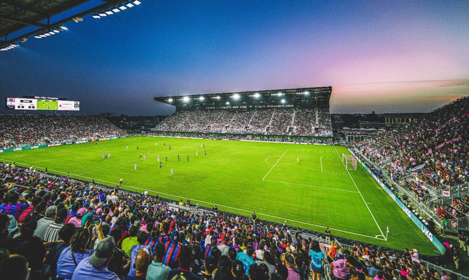 Una vista panorámica de un gran estadio de fútbol moderno al anochecer con reflectores brillantes que iluminan el campo verde donde los jugadores con camisetas azules y rojas se colocan durante un partido rodeados por una multitud de espectadores en varios colores, incluidas camisetas rosas y azules que llenan las gradas.