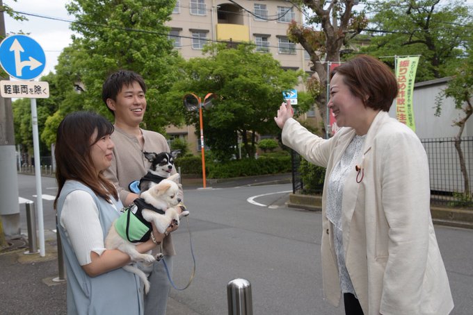 First image shows three people standing on a street near apartment buildings and trees under a partly cloudy sky. A woman in a light blouse holds a small white dog wearing a green vest, another woman in a light top and skirt holds a small black-and-white dog on a leash, and a man in casual clothes smiles while raising his hand. A directional sign and banners are visible nearby. Second image is a newspaper page from Komei Shimbun dated May 10 with Japanese text articles, photos of people at events including outdoor gatherings and group photos, and ads for party subscriptions.