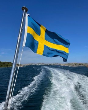 First image shows Swedish flag with blue background and yellow cross waving on a metal pole attached to a boat deck, with blue water surface creating white foam trail behind the boat under clear blue sky and distant rocky shoreline with scattered green vegetation. Second image displays white Bitcoin symbol consisting of B with vertical lines on both sides centered on solid orange background.