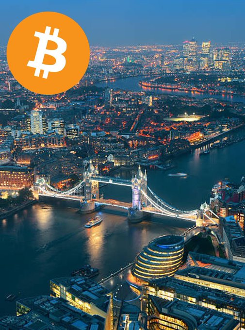 A nighttime aerial view of London cityscape with the River Thames flowing through, illuminated Tower Bridge in the foreground spanning the river with a boat visible on the water, modern buildings and the Gherkin skyscraper in the background, and a large orange Bitcoin symbol overlaid in the upper left corner.