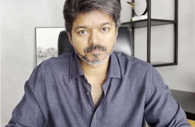 Vijay, a man with gray hair and mustache, sits in a black leather chair wearing a blue shirt, in a modern room with shelves, a small table, and a landscape picture on the wall behind him.