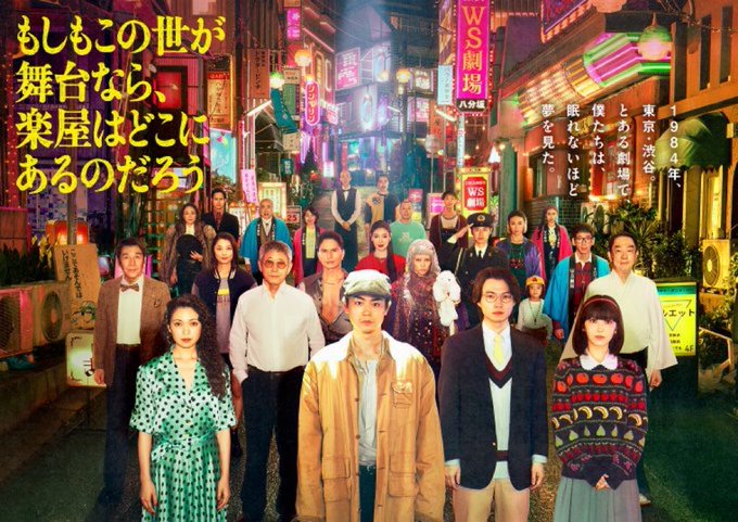 Group promotional poster for a Japanese TV drama set in a neon-lit urban night street scene with Japanese signage including WS and other shop signs. Diverse cast of actors in varied outfits such as suits, dresses, sweaters, and casual wear stand together in the foreground and background, including men in shirts and jackets, women in green dresses and colorful tops, and one in a red hat. The title text in yellow and red kanji reads もしもこの世が舞台なら、楽屋はどこにあるのだろう 絶賛放送中.