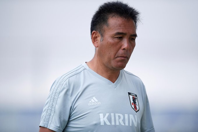 Un hombre asiático de mediana edad con cabello corto y oscuro está de pie de lado con una camiseta de entrenamiento Adidas azul claro con el logotipo del patrocinador Kirin y el emblema de la selección japonesa en el pecho, sobre un fondo exterior borroso que sugiere un campo deportivo.