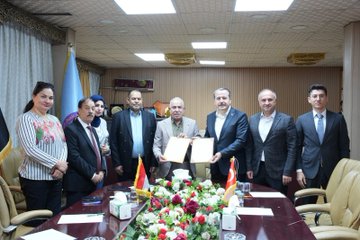First image shows a group of formally dressed men and women standing around a conference table holding documents, with flags of Uzbekistan, Germany, and Turkey on the table adorned with flowers and tissues in a well-lit room. Second image depicts several men in suits seated around a wooden table with food plates, drinks, and portraits of bearded men on the wall in a paneled room. Third image features a group of men in suits standing in front of wooden bookshelves filled with books in a library-like setting. Fourth image displays a modern gray building exterior with a green circular emblem labeled Maarif Schools Baghdad Mansour Campus, and a group of suited men standing in front of the entrance.