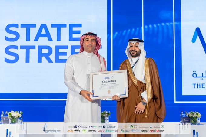 Two men in white thobes and red and white checkered headscarves stand on a stage holding a framed certificate labeled in Arabic and English. The backdrop features a large blue State Street logo with abstract design elements. A table with flowers and sponsor logos including Aramco and HSBC is visible in the foreground. The setting appears to be a formal conference or award event.