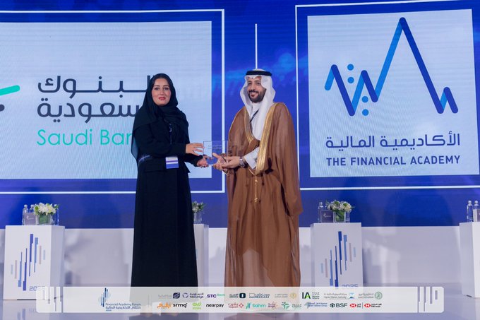 A woman in a black abaya and headscarf stands next to a man in traditional white thobe and red-checkered headscarf on a stage, with the man handing her a trophy. Behind them are blue backdrops with logos of Saudi Banks, The Financial Academy, and sponsors like SNB, SAB, Alinma Bank. Flowers and water bottles are on pedestals beside them.