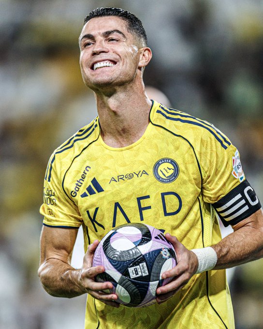 Cristiano Ronaldo smiles upward while holding a purple and white soccer ball in a stadium crowd. He wears a yellow Al-Nassr jersey with Adidas and KAFD logos on the chest and black armbands. The jersey features blue accents and sponsor patches. His hair is short and dark, and he has a beard.