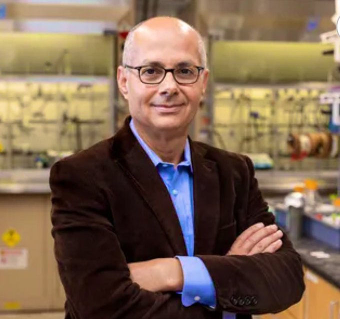 A bald man with glasses wearing a brown blazer and blue shirt stands with arms crossed in a laboratory setting surrounded by scientific equipment shelves and yellow safety signs.