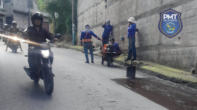 Several police officers in blue uniforms and helmets stand near motorcycles on a wet street, one holding a phone, while workers in vests and hats use tools and a cart to fill potholes beside a gray concrete wall with the P M T Villa Nueva shield emblem, surrounded by urban greenery and a barrel.