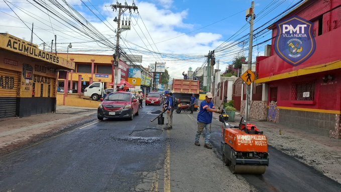 Street scene in Villa Nueva zona 1 with workers in blue uniforms patching asphalt potholes using an orange steamroller machine and brooms on 3a calle and 10a avenida. Red and yellow buildings with PMT logos line the road, several vehicles including a white van and red car are parked nearby, utility poles and traffic signs visible under a partly cloudy sky.