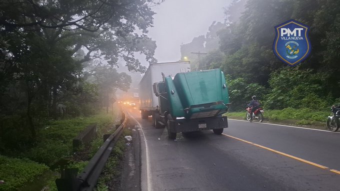 A green truck trailer with mechanical issues is stopped on a misty uphill road surrounded by dense green trees and vegetation. Police motorcycles are positioned nearby, and a PMT Villa Nueva badge is visible in the upper right. The scene includes a line of vehicles with headlights on in the fog, and a road barrier on the side.