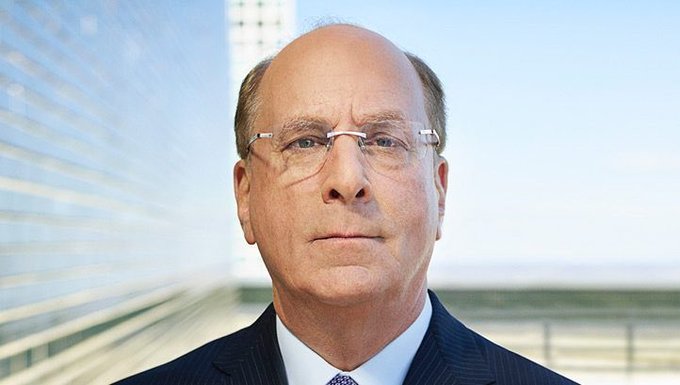 Man in suit and glasses stands in front of modern glass building exterior under blue sky. Prominent white Bitcoin symbol displayed on orange background.