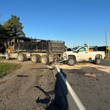 First image displays a white pickup truck with front-end damage positioned beside a black dump truck on a rural roadside under clear skies with utility poles and grassy fields in background showing collision aftermath. Second image shows a dump truck with side damage next to a white pickup truck on roadside with dirt ground and trees under blue sky depicting vehicle crash scene.