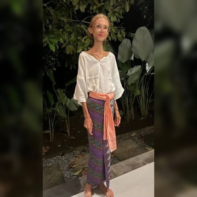 Kadın açık havada yeşil yapraklı tropik bitkilerin önünde ayakta duruyor. Beyaz bol kollu bluz ve mor turuncu desenli uzun etek giymiş. Bacakları zayıf görünüyor ve yalınayak.