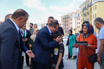 First image shows a group of men in suits and uniforms standing at a podium with a Turkish flag banner, clapping, in front of modern apartment buildings under a clear sky with a drone flying above. Second image depicts officials including men in suits and caps gathered around a podium, one speaking into a microphone, with buildings and a yellow banner in the background. Third image features a man in a blue suit hugging a child while surrounded by women in headscarves and modest clothing, holding a tablet, on a paved area with apartment buildings. Fourth image displays a large group of men and women in formal attire posing in front of a yellow building entrance labeled E12 with a blue sign, some holding a baby, near benches.